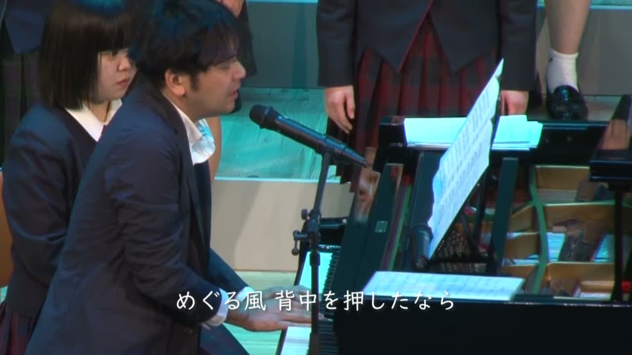ステージでピアノを弾きながらマイクに向かって歌う弓削田健介（「越えてゆけ」暁学園合唱部とのコラボより）