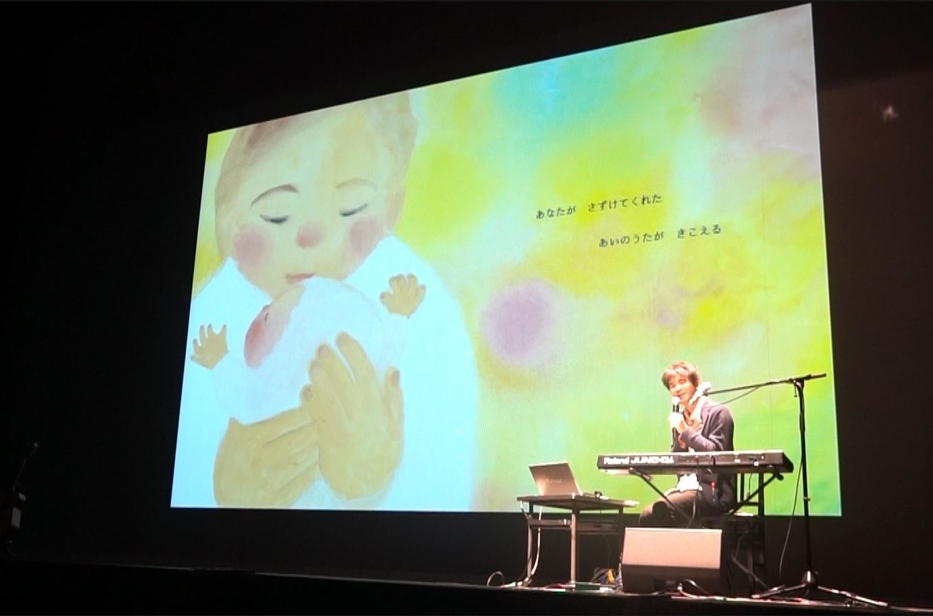 暗いホールの大きなスクリーンに絵本の絵が映り、その前で歌う弓削田健介